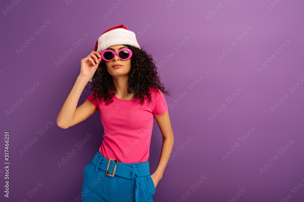 stylish mixed race girl in santa hat touching glasses while holding hand in pocket on purple background stylish mixed race girl in santa hat touching glasses while holding hand in pocket on purple background