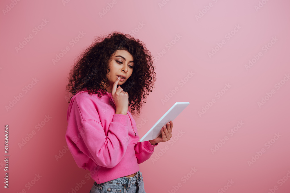 thoughtful bi-racial girl touching lip while using digital tablet on pink background thoughtful bi-racial girl touching lip while using digital tablet on pink background