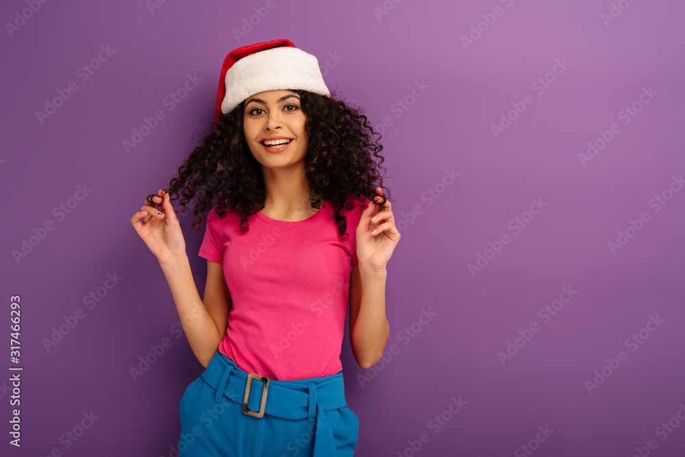 attractive mixed race girl in santa hat touching hair while smiling at camera on purple background attractive mixed race girl in santa hat touching hair while smiling at camera on purple background