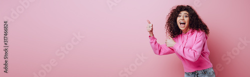 panoramic shot of excited bi-racial girl showing thumbs up on pink background