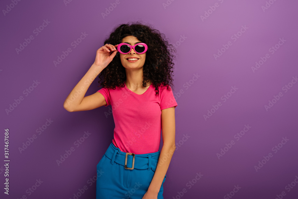 cheerful bi-racial girl touching sunglasses while looking at camera on purple background