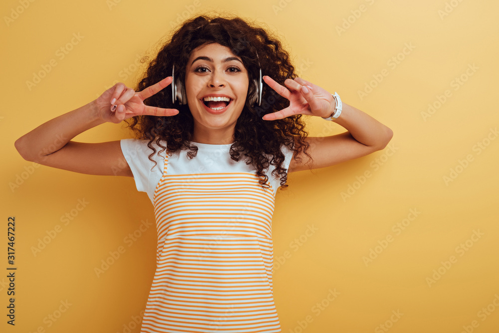 cheerful mixed race girl in wireless headphones showing victory gesture on yellow background cheerful mixed race girl in wireless headphones showing victory gesture on yellow background