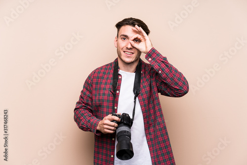 Young caucasian photographer man isolated excited keeping ok gesture on eye.