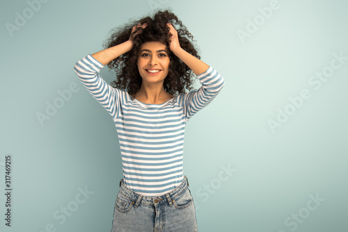 happy mixed race girl touching hair while smiling at camera on grey background