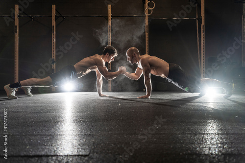 Athletic sportsmen doing push ups together and holding hands in crossfit gym