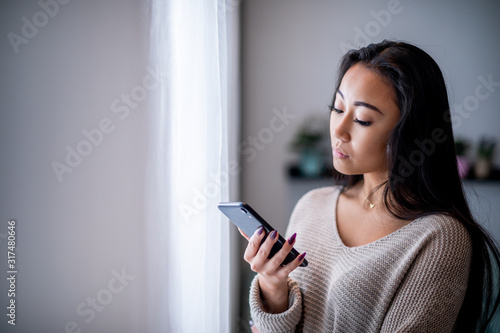 Upset asian woman standing near window with smartphone and reading message