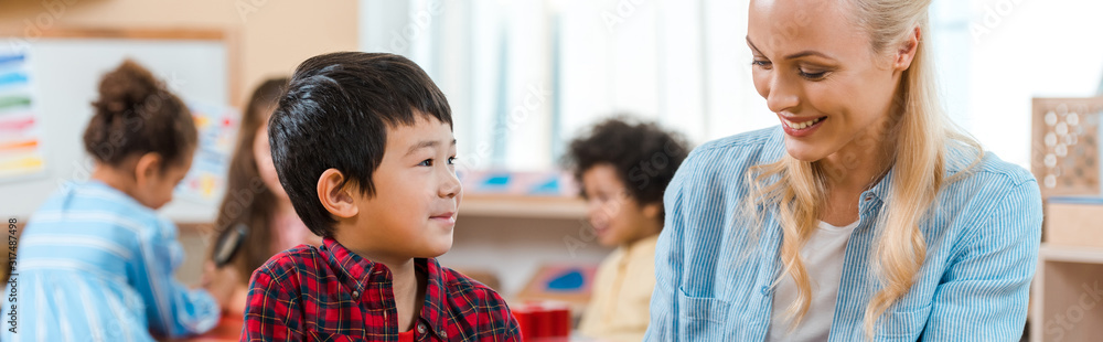 Panoramic shot of smiling teacher and kid with children at background in montessori school Panoramic shot of smiling teacher and kid with children at background in montessori school