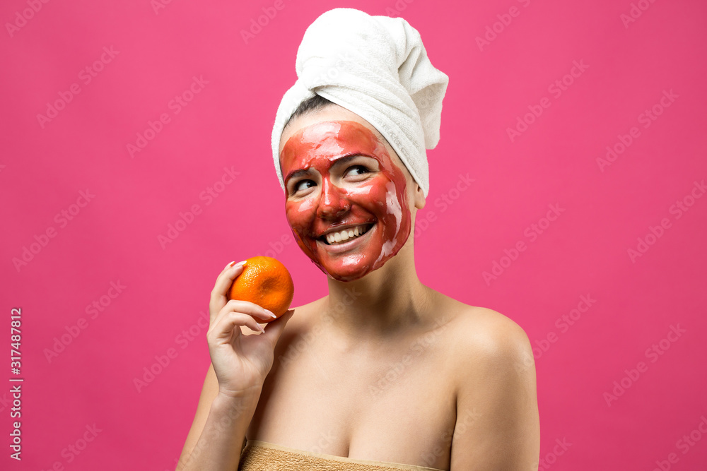 Beauty portrait of woman in white towel on head with red nourishing mask on face. Skincare cleansing eco organic cosmetic spa relax concept. A girl stands with her back holding an orange mandarin. Beauty portrait of woman in white towel on head with red nourishing mask on face. Skincare cleansing eco organic cosmetic spa relax concept. A girl stands with her back holding an orange mandarin.