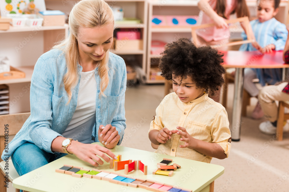 Fototapeta premium Selektywne skupienie się na dzieciaku grającym w drewnianą grę przez nauczyciela z dziećmi w tle w szkole montessori