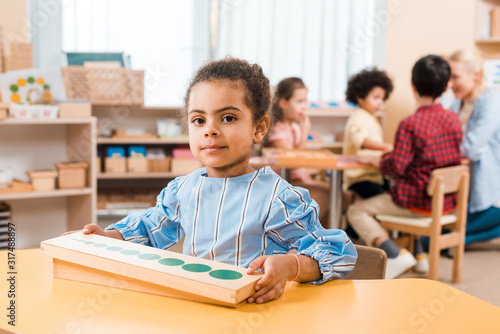 Selective focus of kid with wooden game looking at camera and teacher with children at background in montessori school