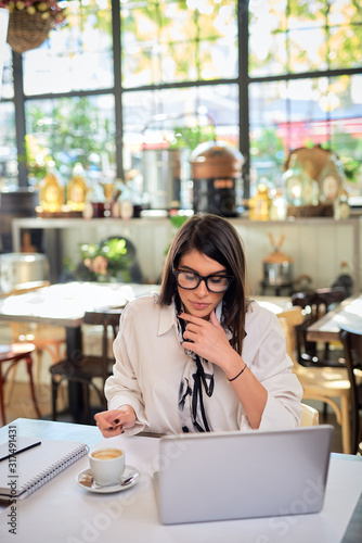 Attractive smiling caucasian female lawyer with eyeglasses dressed in shirt sitting in cafe and using laptop for work.