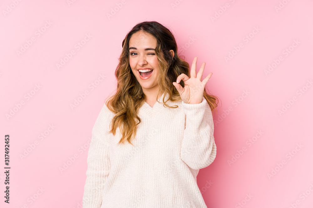 Young curvy woman posing in a pink background isolated winks an eye and holds an okay gesture with hand. Young curvy woman posing in a pink background isolated winks an eye and holds an okay gesture with hand.