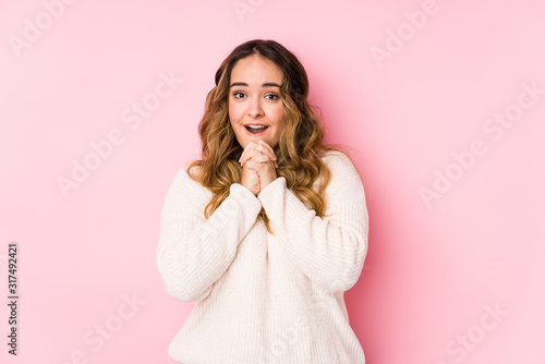 Young curvy woman posing in a pink background isolated praying for luck, amazed and opening mouth looking to front.