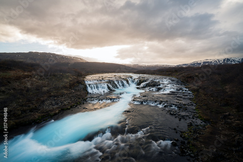 Bruarfoss waterfall in Icelandic scenery during sunset and cloudy sky. Turquoise cascade and golden warm light with snow covered mountains in the background.