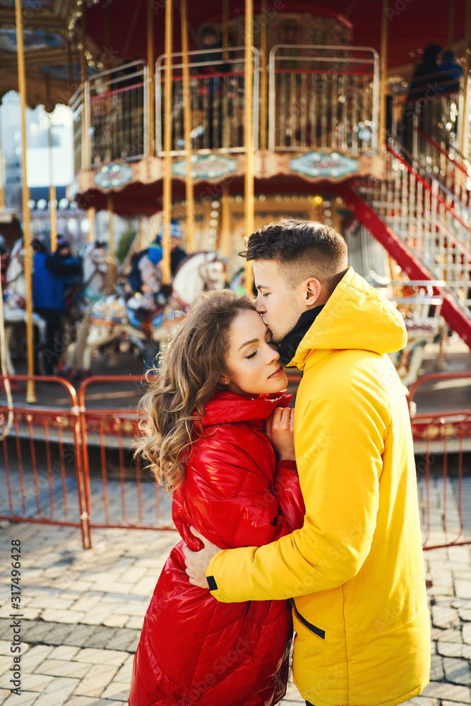 young romantic couple in love wearing in bright yellow and red down jackets hugging on french carousel background.