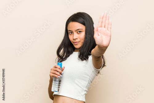 Young asian woman holding a water bottle standing with outstretched hand showing stop sign, preventing you.