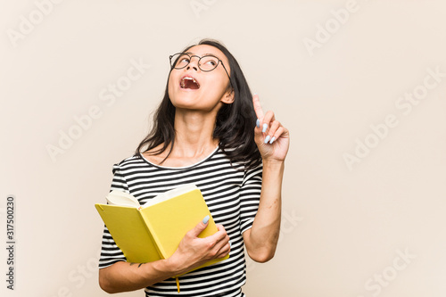 Young asian woman student holding a book pointing upside with opened mouth.