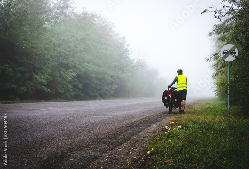 Cyclist with reflective vest and lighs on is walking down the road to the fog in nature. Bicycle touring in bad  weather conditions and bad visability. photo