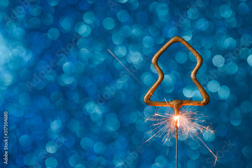 Burning sparkler in christmas tree shape on blue glitter background with copy-space.