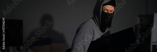 Tableau sur toile Portrait of male thug standing in dark room and holding expensive televisor from popular company