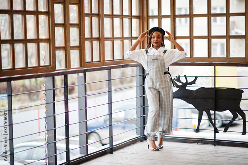 African american woman in overalls and beret posed in outdoor terrace with christmas decorations garland and deer.