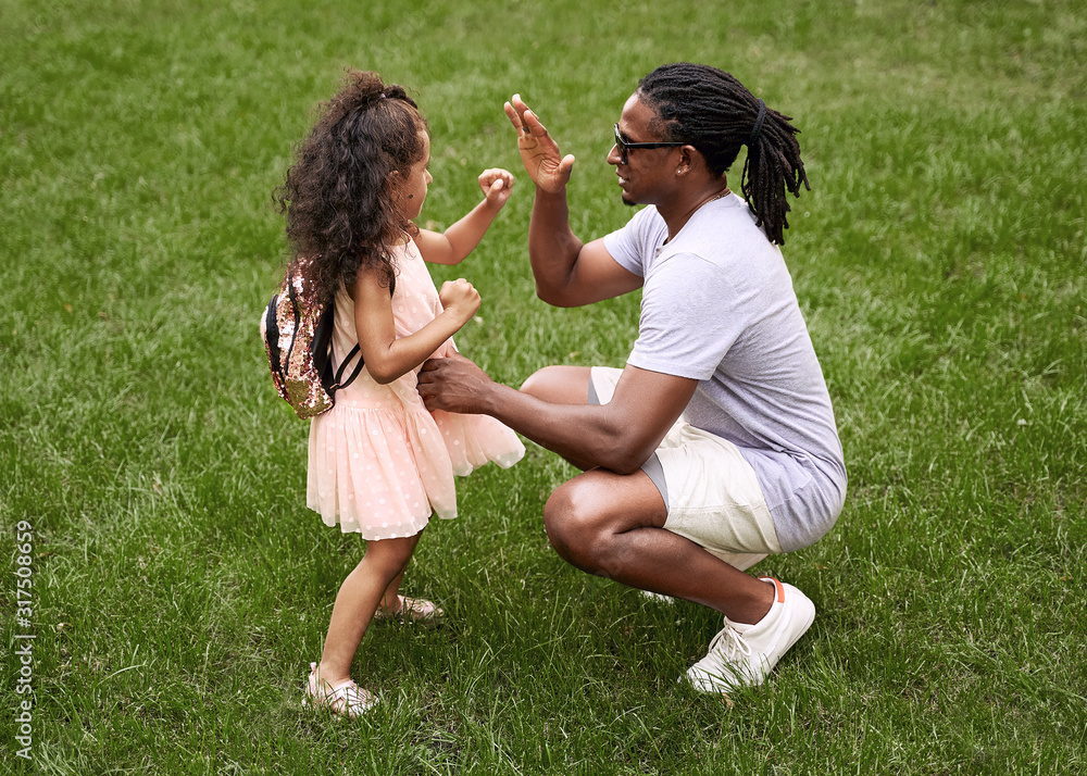 Happy multiethnic family. Fit black father teaching his adorable cute mixed race daughter in pink dress how to fight on the green grass lawn. Boxing karate or other martial arts outdoor fitness class.