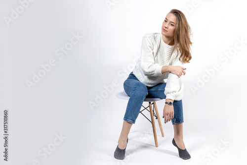 attractive young girl in jeans and a white sweater sits on a chair on a white background, copy space