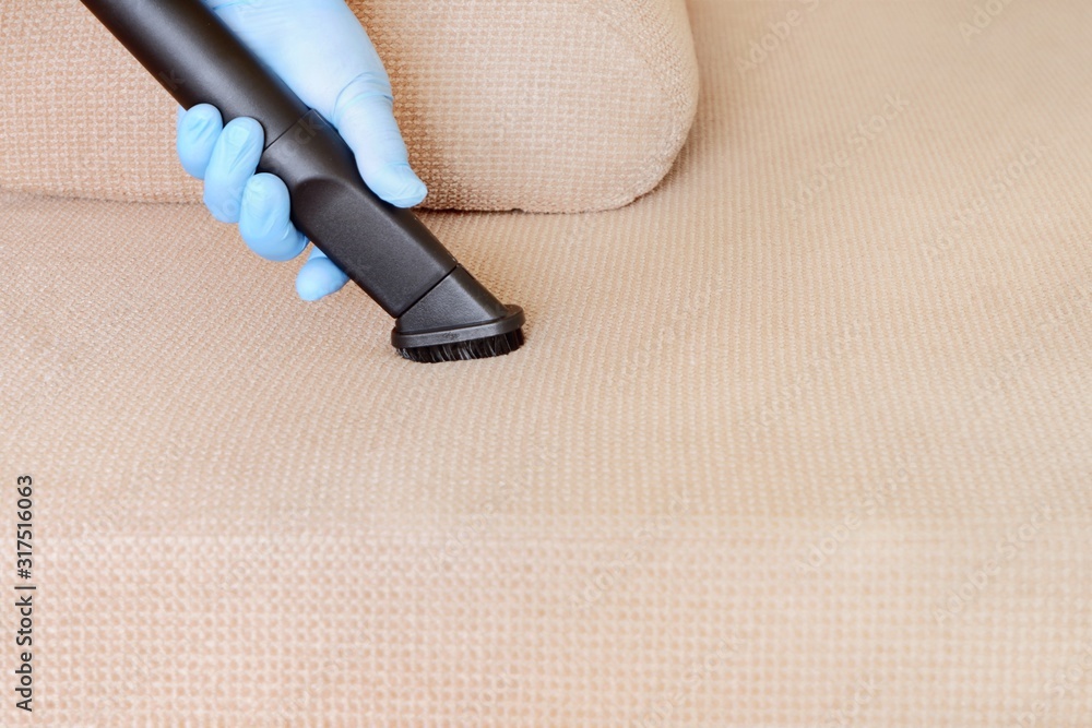 Fototapeta premium A female hand in a glove vacuums the surface of the sofa. The concept of home cleaning, housekeeping, early spring regular cleanup. Close-up, selective focus, copy space for text.