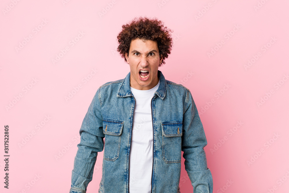 Curly mature man wearing a denim jacket against pink background screaming very angry and aggressive. Curly mature man wearing a denim jacket against pink background screaming very angry and aggressive.