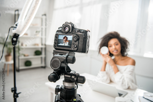 selective focus of digital camera with attractive african american influencer in braces holding container with cosmetic cream on display