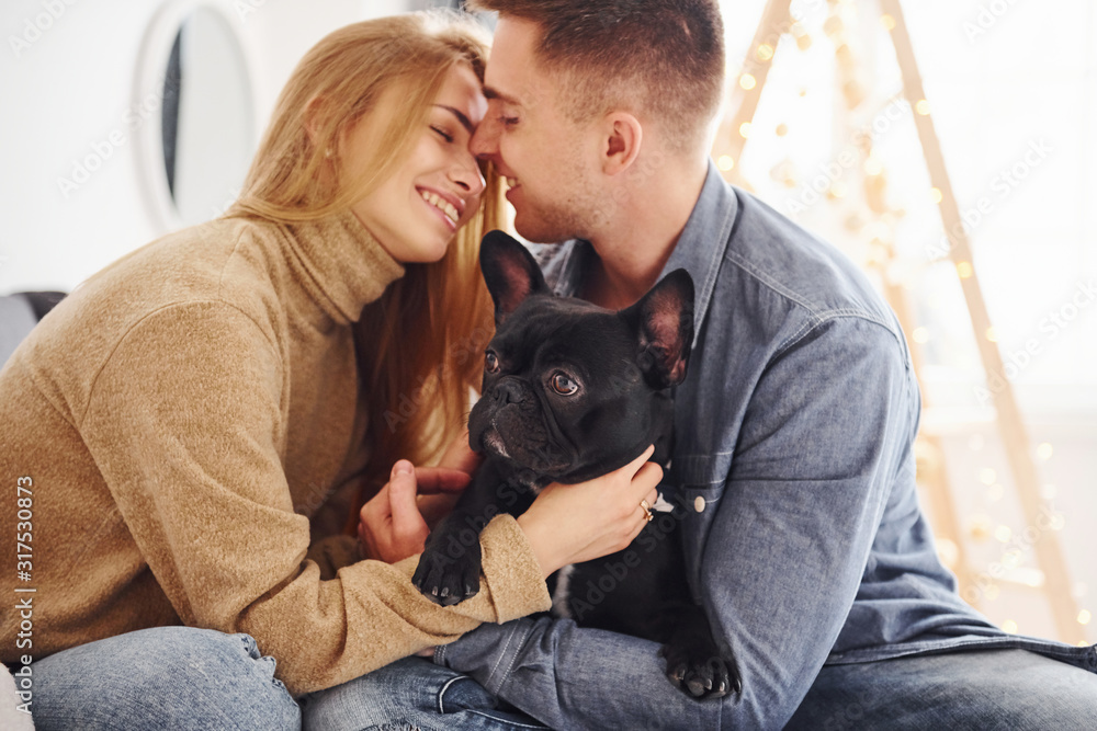 Happy young modern couple sitting on the sofa at home with christmas tree with their cute dog