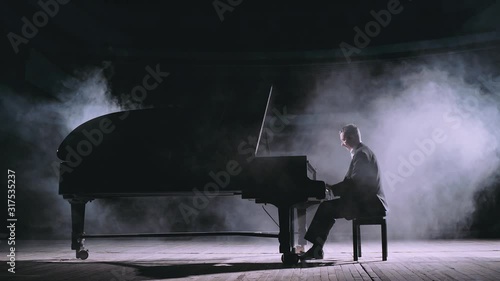 Professional pianist in dark suit playing on a grand piano on big stage in concert hall with dimmed light in the smoke