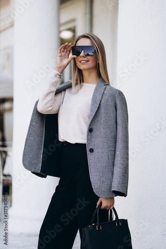 Outdoor photo of blonde lady posing on architecture background in autumn day.Fashion street style portrait. wearing dark casual trousers and creamy sweater and sunglasses.Fashion concept.