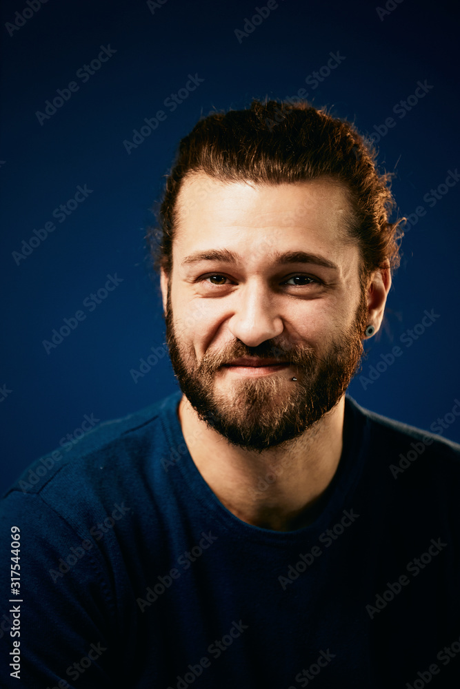 Young handsome smiling bearded caucasian hipster in blue sweater standing in front of blue background and looking at camera.