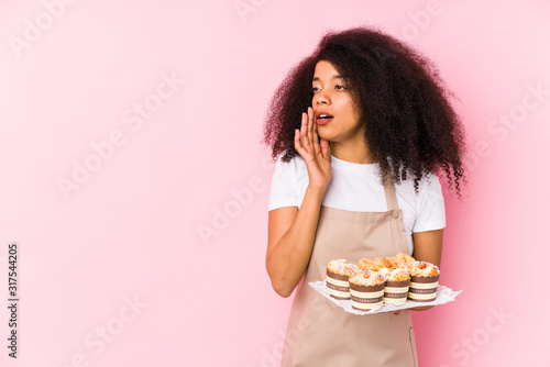 Young afro pastry maker woman holding a cupcakes isolatedYoung afro baker woman is saying a secret hot braking news and looking aside