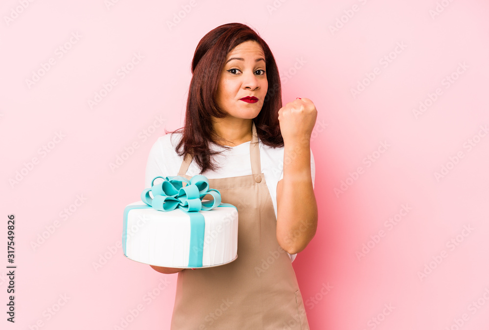 Middle age latin woman holding a cake isolated on a pink background showing fist to camera, aggressive facial expression. Middle age latin woman holding a cake isolated on a pink background showing fist to camera, aggressive facial expression.