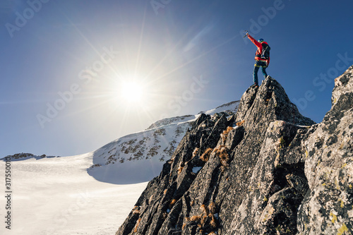 Climber or alpinist at the top of a mountain. A success of mountaineer reaching the summit. Outdoor adventure sports in winter alpine moutain landscape. Sunny day and a climber on a top of a peak.