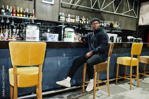 Handsome african american man posing inside night club in black hat, sitting on bar counter.