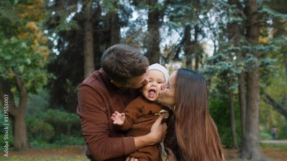 Young mother and father holding cute little child in their hands and kissing him from both sides outdoors. Cheerful child is smiling. Happy family having fun in park. Young mother and father holding cute little child in their hands and kissing him from both sides outdoors. Cheerful child is smiling. Happy family having fun in park.