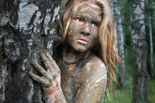 A girl with a mud mask peeps out from behind a birch trunk at an ecological resort. Beautiful blonde