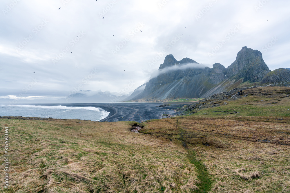 View over Eystrahorn mountain chain and Hvalnes beach next to the Ring road in Iceland. Seagulls flying overhead. Foot path leading towards the peak with foggy ring around it.