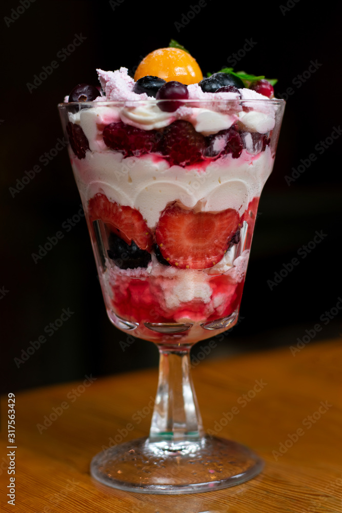 Colorful trifle dessert with fizalis, strawberries, blueberries, jam and whipped cream on high translucent glass on wooden table, vertical photography in low-key lightning Colorful trifle dessert with fizalis, strawberries, blueberries, jam and whipped cream on high translucent glass on wooden table, vertical photography in low-key lightning