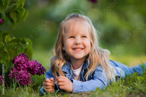 little blonde girl lies near the lilac flowers on the green grass and smiles © КРИСТИНА Игумнова