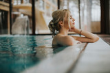 Young woman with long blonde hair, pulled back in a simple bun, sits at the edge of a pristine indoor pool, eyes closed and a serene expression on her face