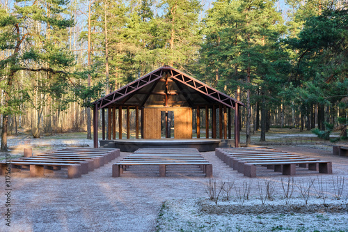 Wooden house stage for free musicians open air performances in pine forest in sunlight, in front of stage many benches covered with snow. Raising  culture of people, music for all, art theater in park