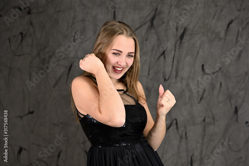 A photo of a pretty smiling girl with long hair and excellent make-up in a black dress stands in different poses. Universal concept of horizontal female portrait on gray alternative background.