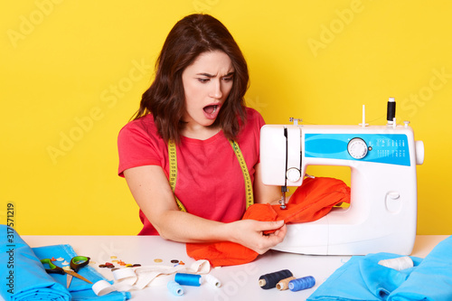 Picture of shocked desperate young inexperienced seamstress working with red fabric, having troubles with sewing, having unpleasant facial expression, opening her mouth widely. Work concept.