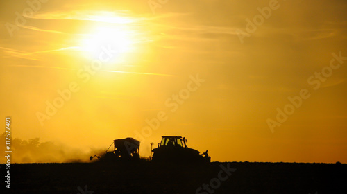 Silhouette of a tractor sowing seeds in a field in a cloud of dust against the background of the setting sun.