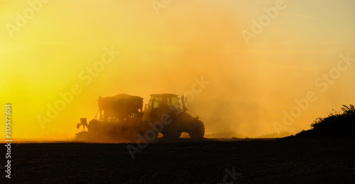 Silhouette of a tractor sowing seeds in a field in a cloud of dust against the background of the setting sun.