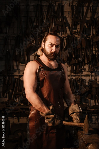 Brutal muscular blacksmith in a dark workshop forges iron
